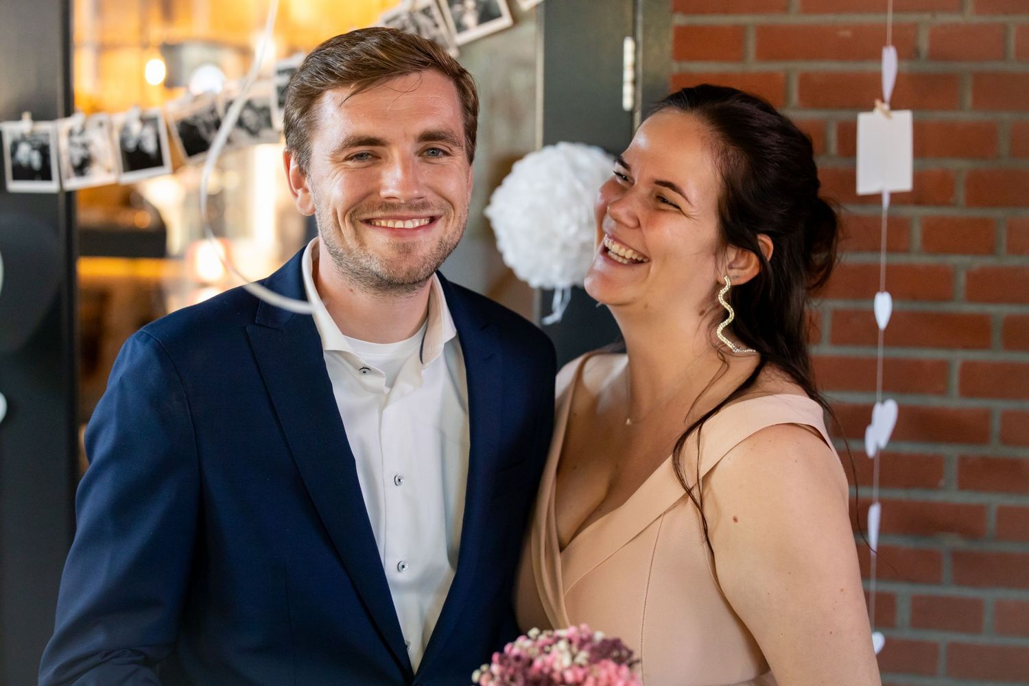 Happy couple smiling at a wedding, man in blue suit, woman in beige dress holding a bouquet, festive decorations behind.