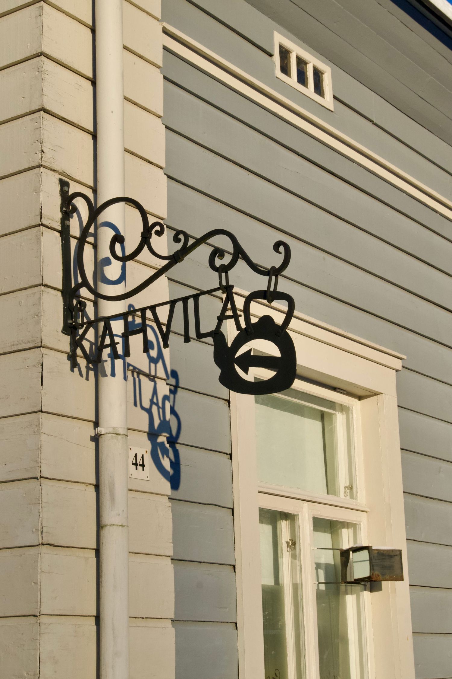 Ornate wrought iron coffee shop sign with teapot silhouette mounted on corner of gray wooden building, number 44.
Reizen - Lynn - Helsinki