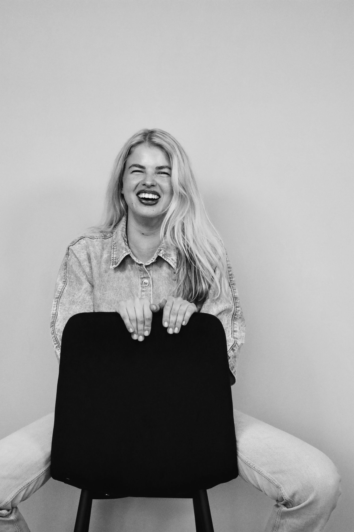Black and white photo of a laughing blonde woman in a denim shirt sitting backwards on a chair against a light background.
Tess - Soloshoot