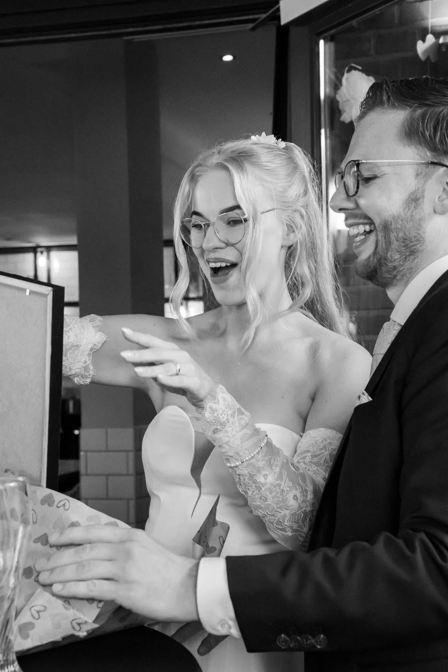 Black and white photo of a laughing bride and groom in glasses receiving a gift at their wedding reception.