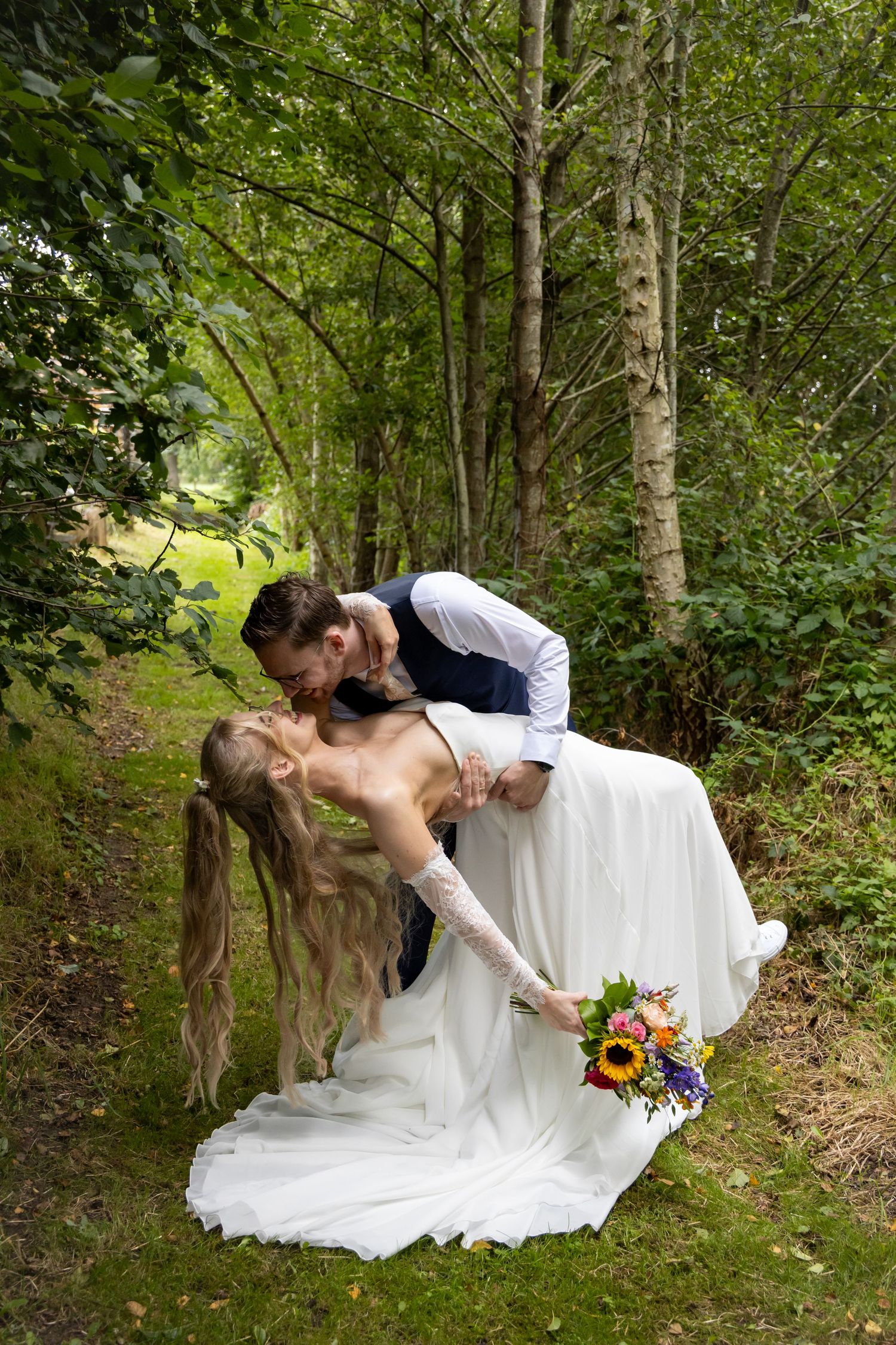 Groom dipping bride for a romantic kiss in a woodland setting, bride holding a colorful bouquet.