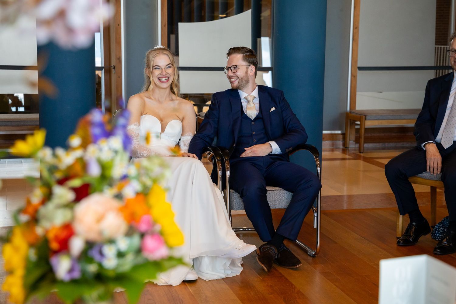 Bride and groom sitting together smiling during wedding ceremony with colorful floral arrangement in foreground.