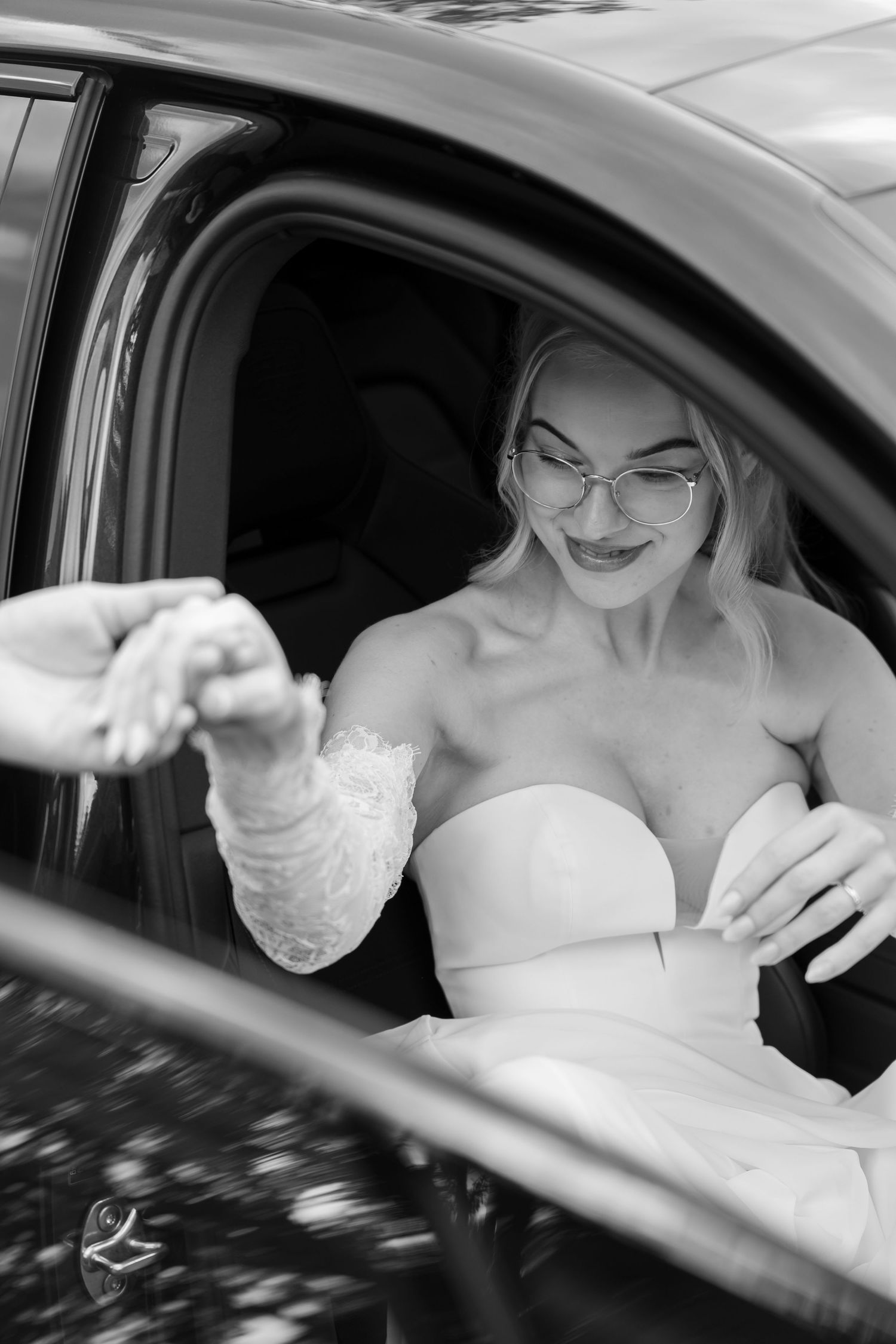 Blonde bride in strapless white gown wearing glasses sits in car, reaching out to hold hand in black and white photo.
