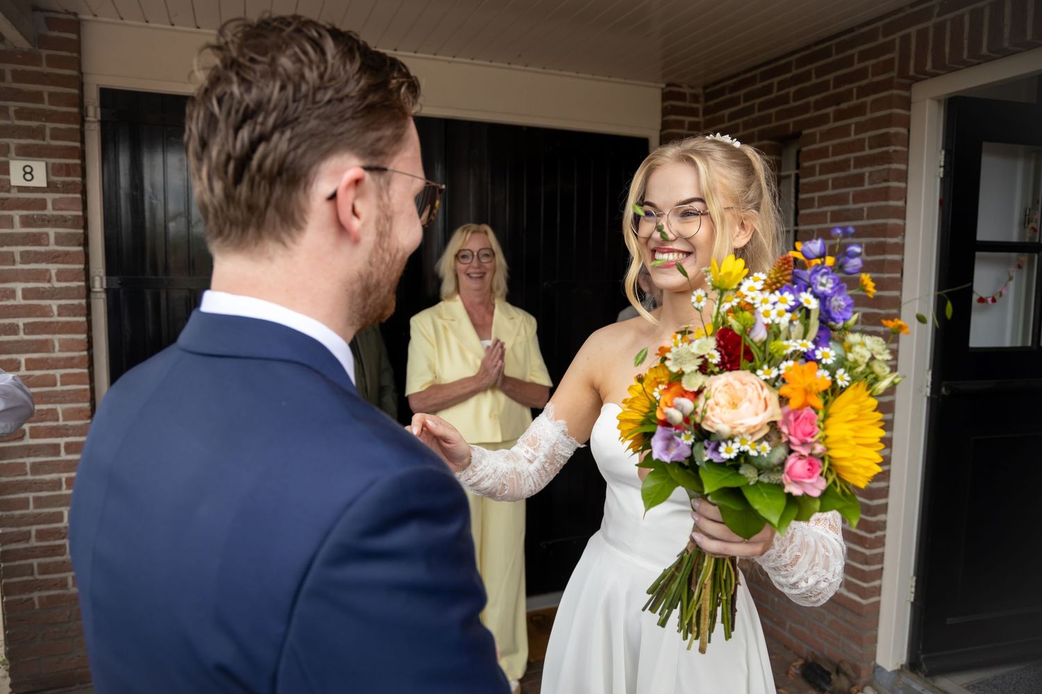 Bride in white dress holds colorful bouquet, smiling at groom in blue suit, with woman in background.