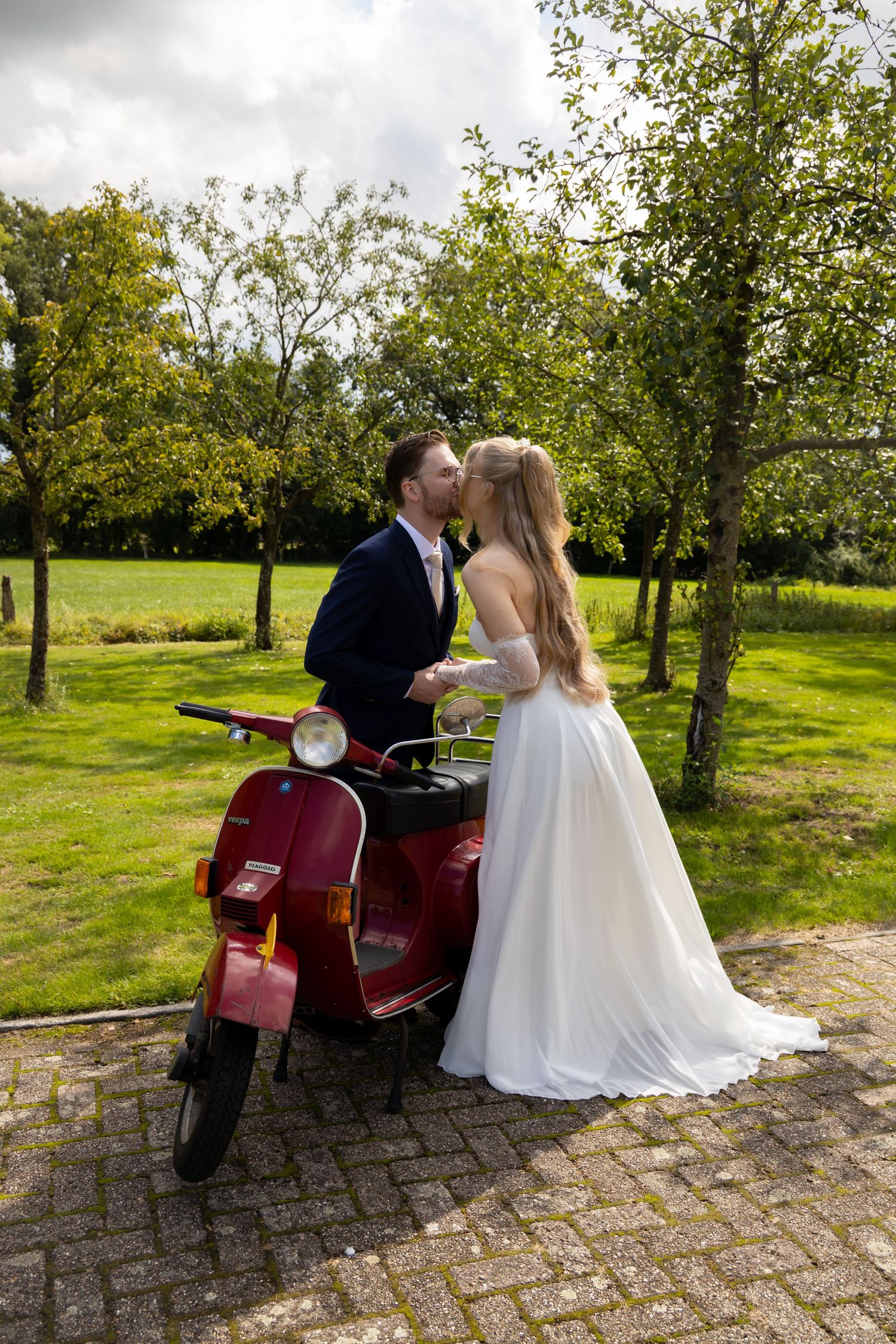 Bride and groom kissing beside a red Vespa scooter on a sunny day in a lush green garden.