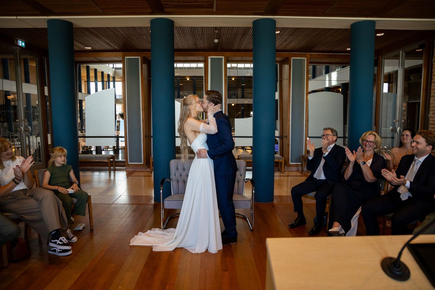 Bride and groom share first kiss at wedding ceremony while guests applaud in elegant venue with teal columns.