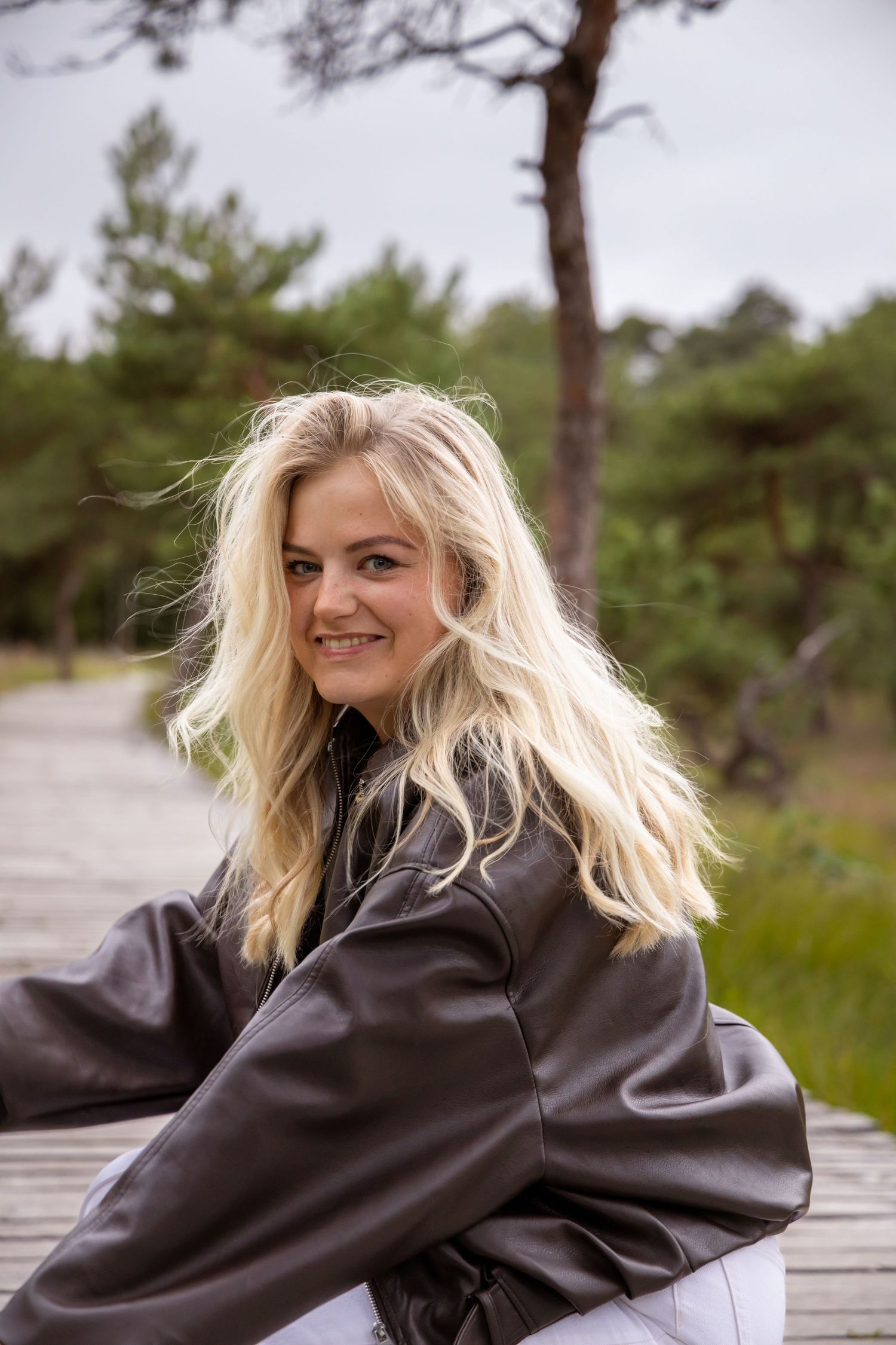Smiling blonde woman in brown leather jacket sitting on wooden boardwalk surrounded by green trees outdoors.