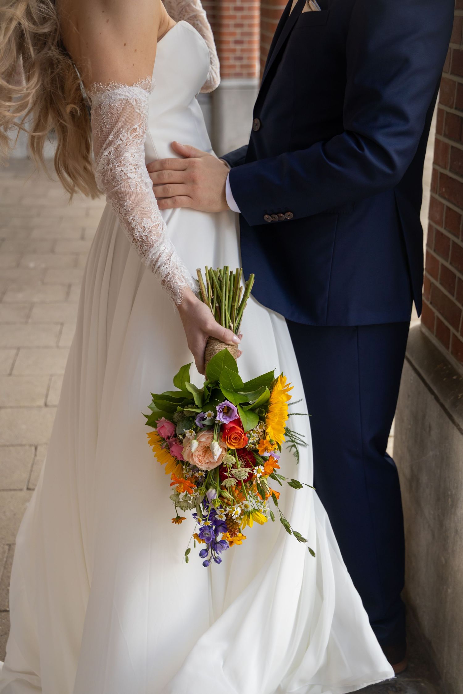 Bride in lace wedding gown holds colorful bouquet while groom in navy suit embraces her waist outdoors.