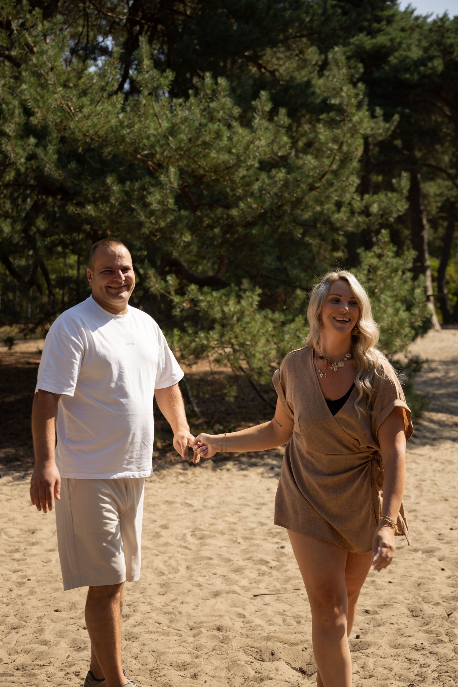 A smiling couple holding hands while walking on a sandy beach path surrounded by green pine trees.
Familieshoot - Koppelshoot