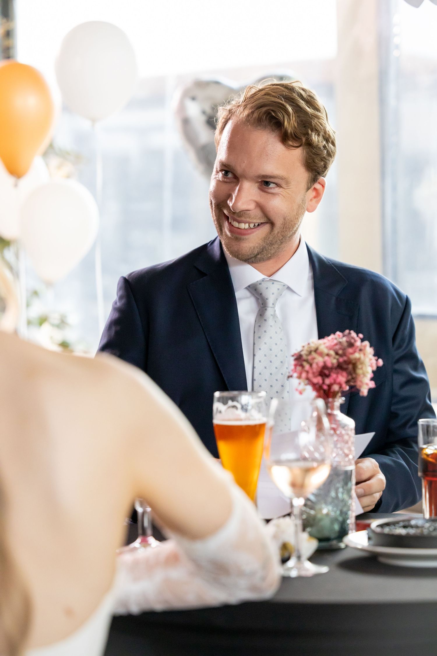 Smiling man in navy suit sits at wedding reception table with beer, beside bride in white lace dress and balloon decorations.