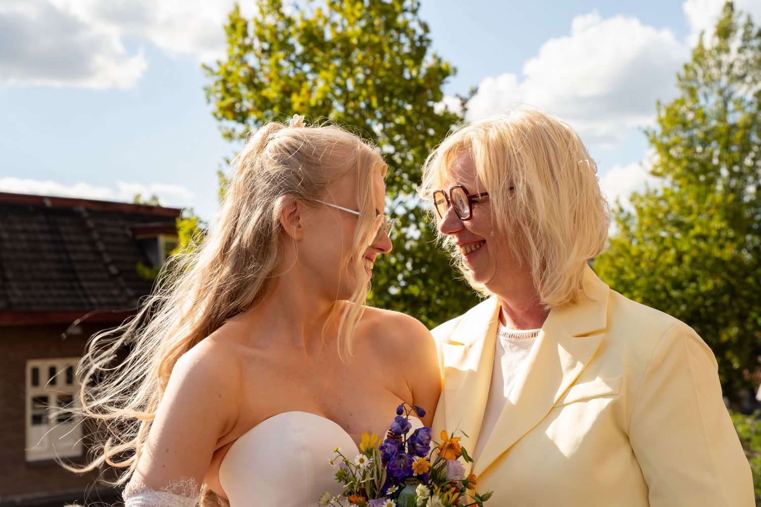 Two blonde women in wedding attire smile at each other outdoors, one holding a colorful bouquet on a sunny day.