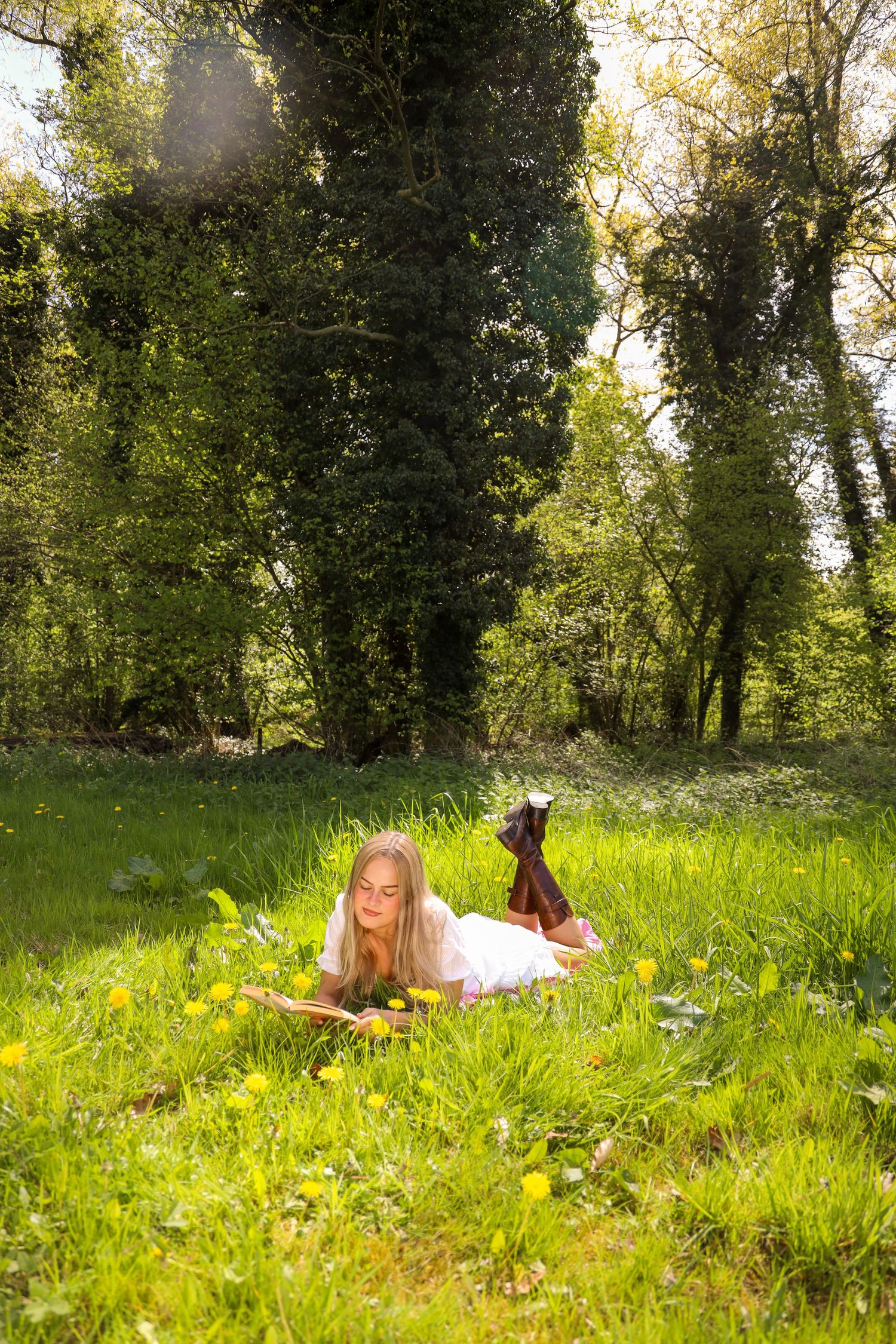 Young woman in white dress reading a book while lying on green grass in a sunny forest clearing with yellow wildflowers.
Romy - Soloshoot