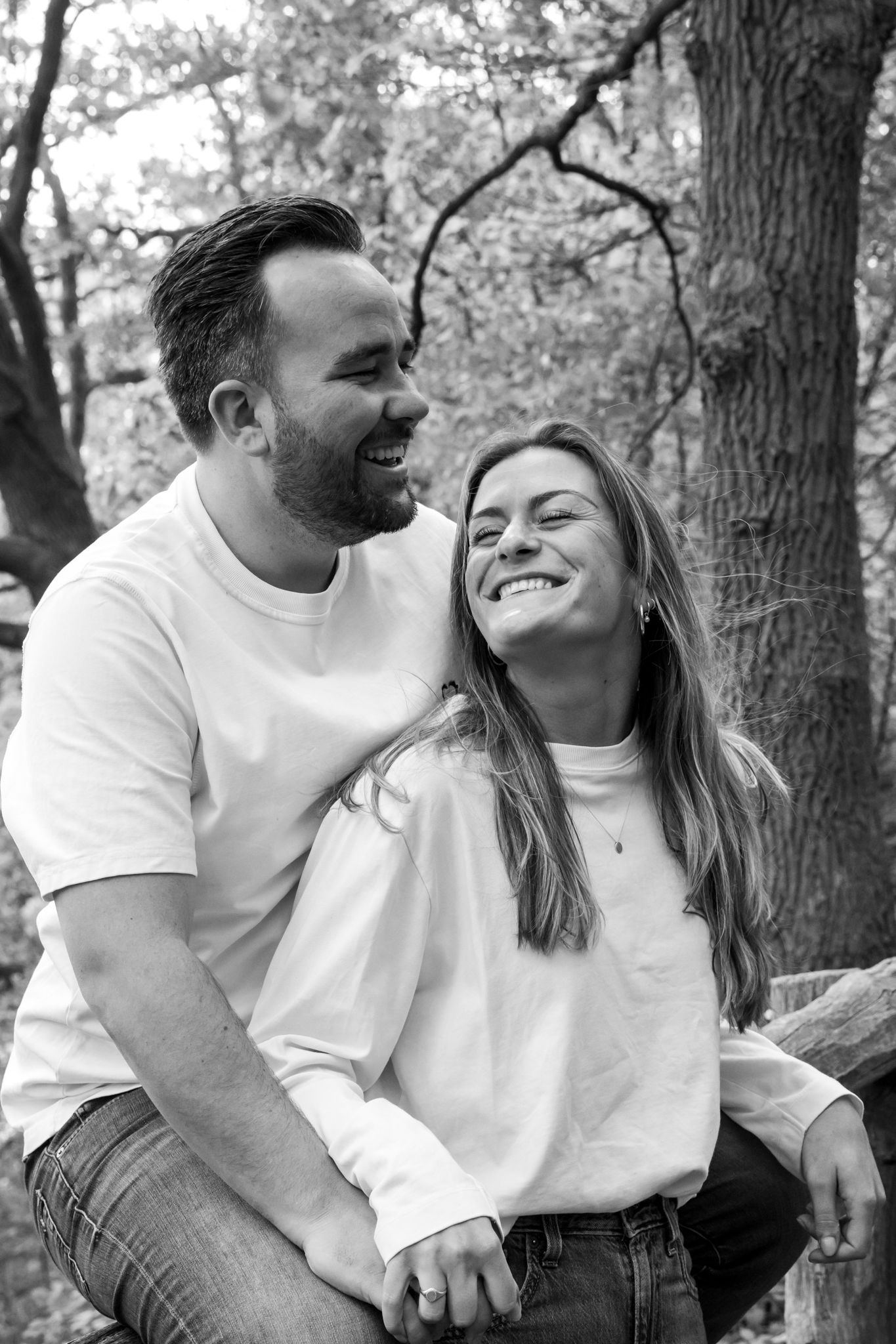 Black and white photo of a smiling couple embracing outdoors among trees, wearing casual white tops.
Fleur en Timo - Koppelshoot