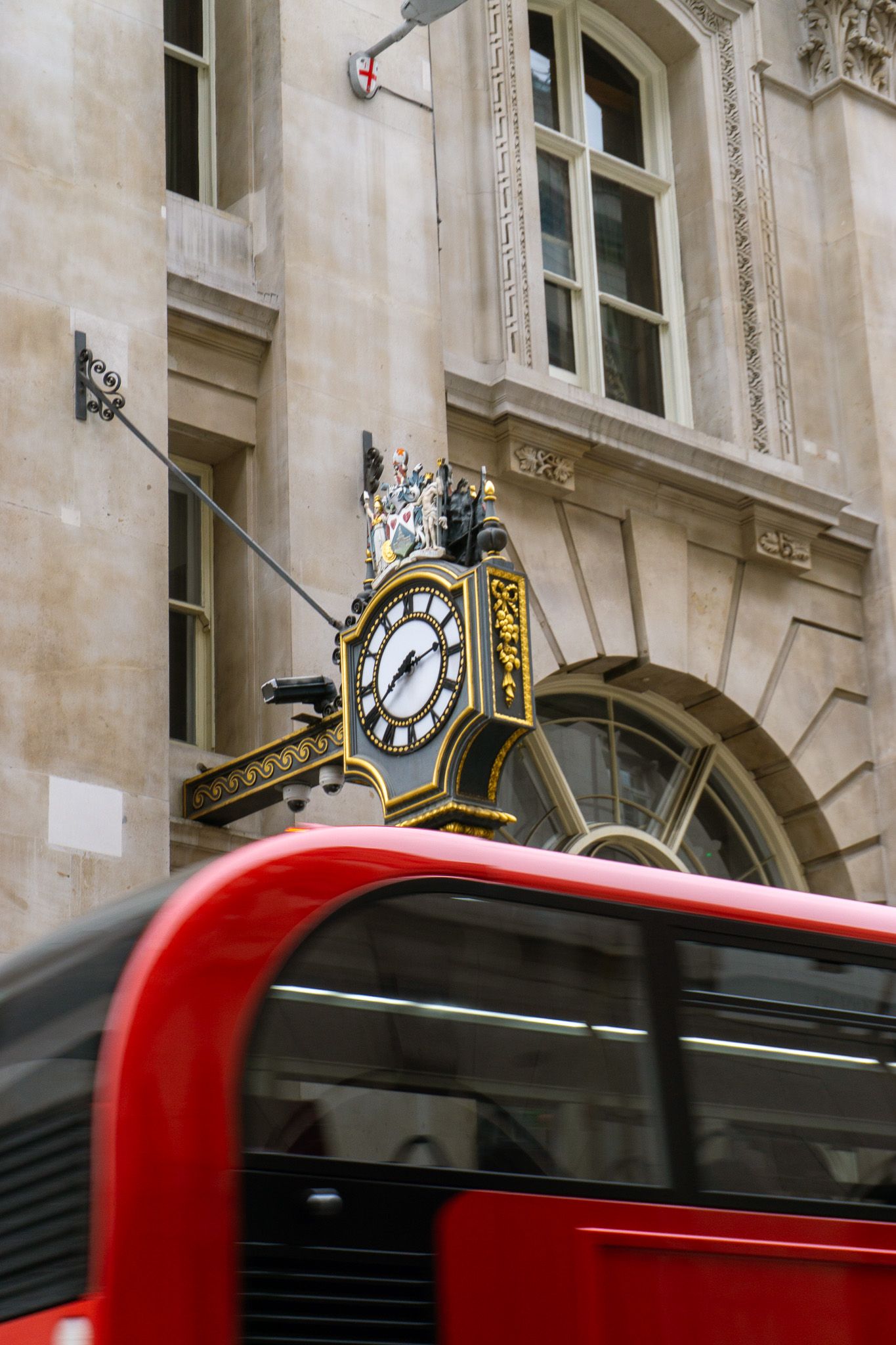 Ornate black and gold clock on a London building facade with a red double-decker bus passing below.
Reizen - Lynn - Londen