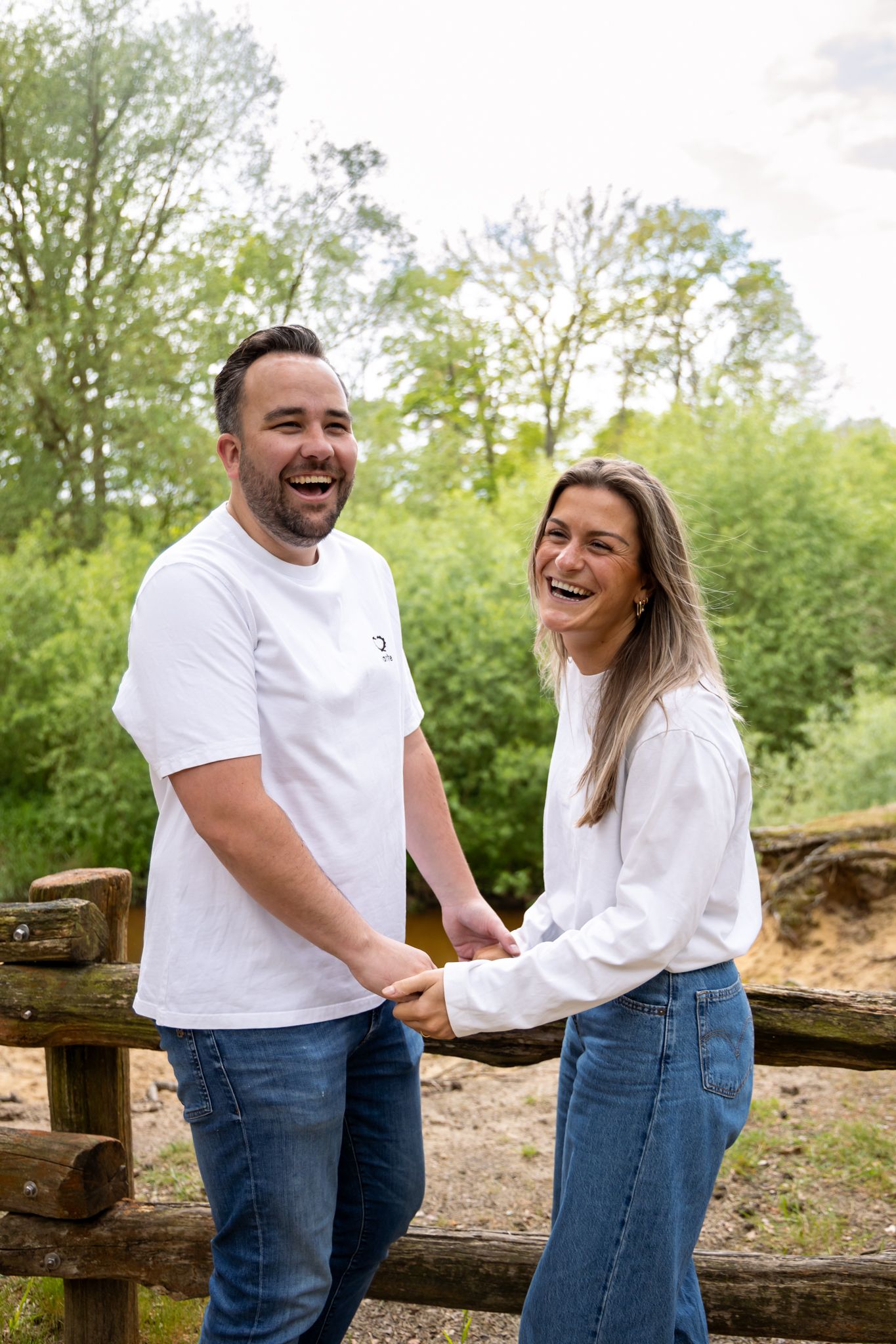 A laughing couple in matching white tops and jeans hold hands by a wooden fence with green trees behind them.
Fleur en Timo - Koppelshoot