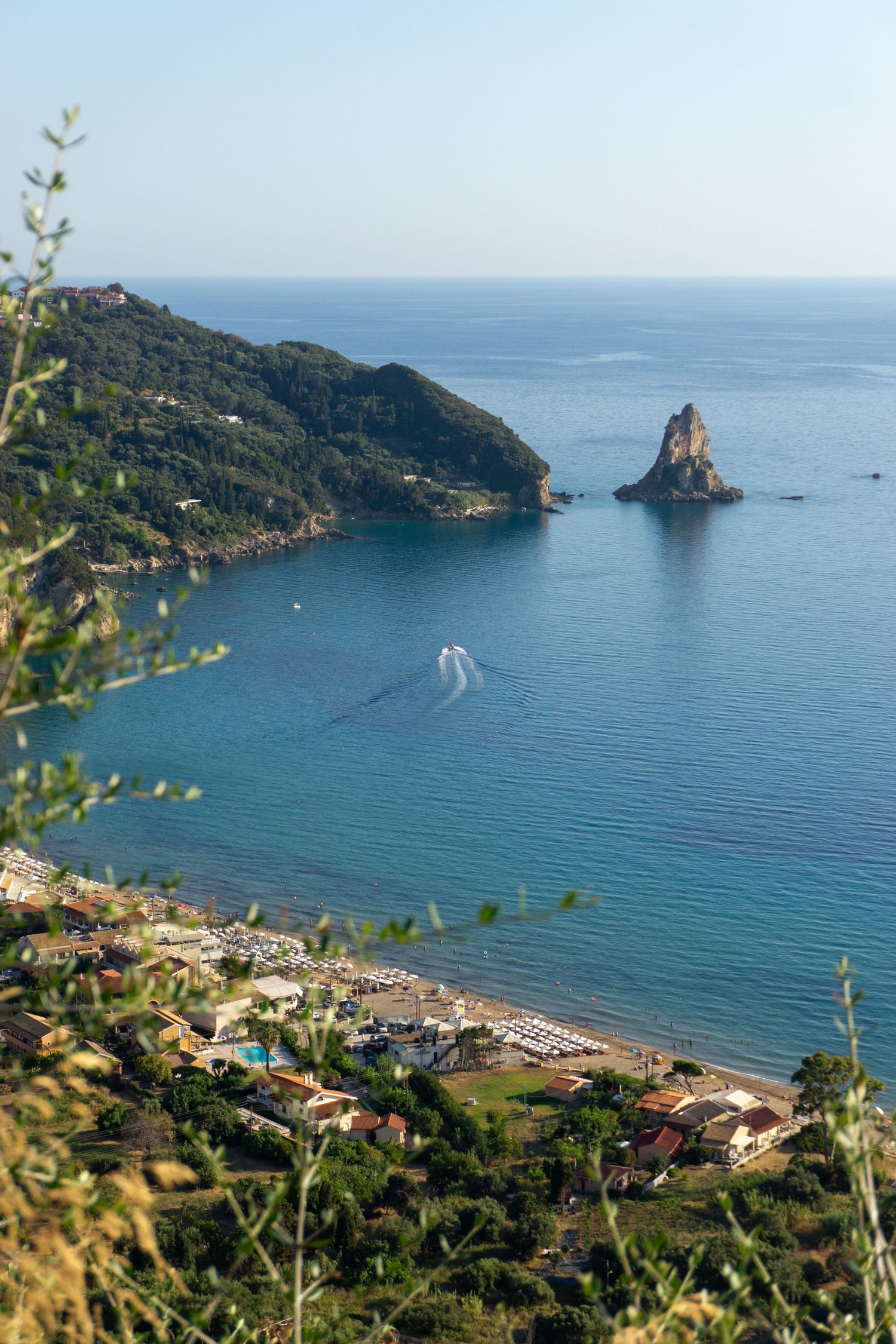 Aerial view of a turquoise Mediterranean bay with a rocky outcrop, sandy beach, and lush green hillside in Corfu, Greece.