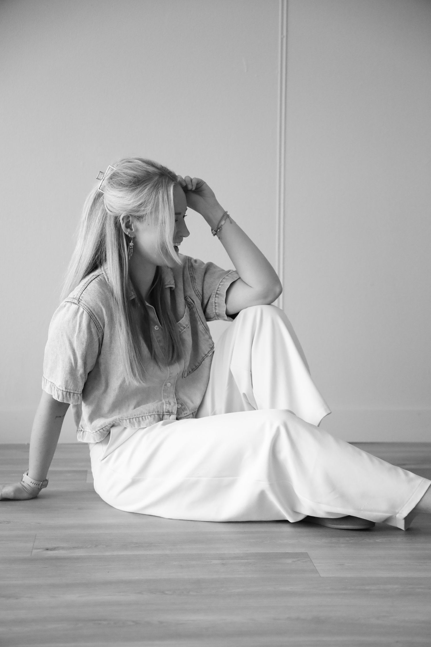 Black and white photo of a blonde woman sitting on a wooden floor in white pants and denim shirt, hand in hair.