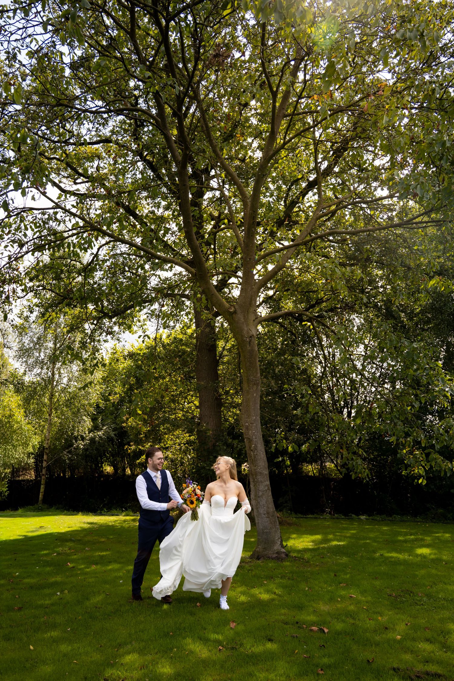 Bride and groom walking hand in hand on lush green lawn beneath a large tree on their wedding day.