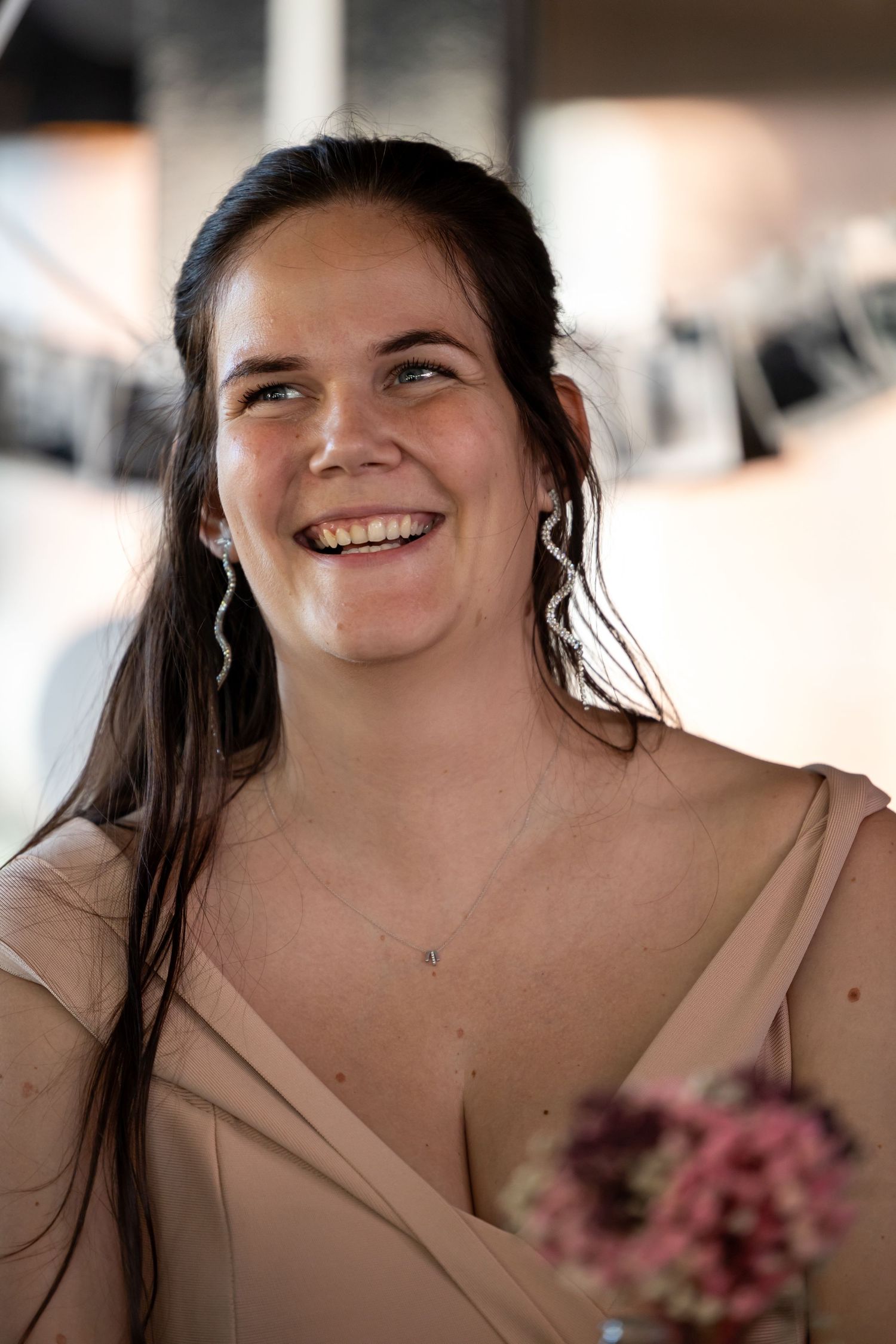 Smiling brunette woman in a beige off-shoulder dress holding pink flowers, wearing silver earrings and necklace.