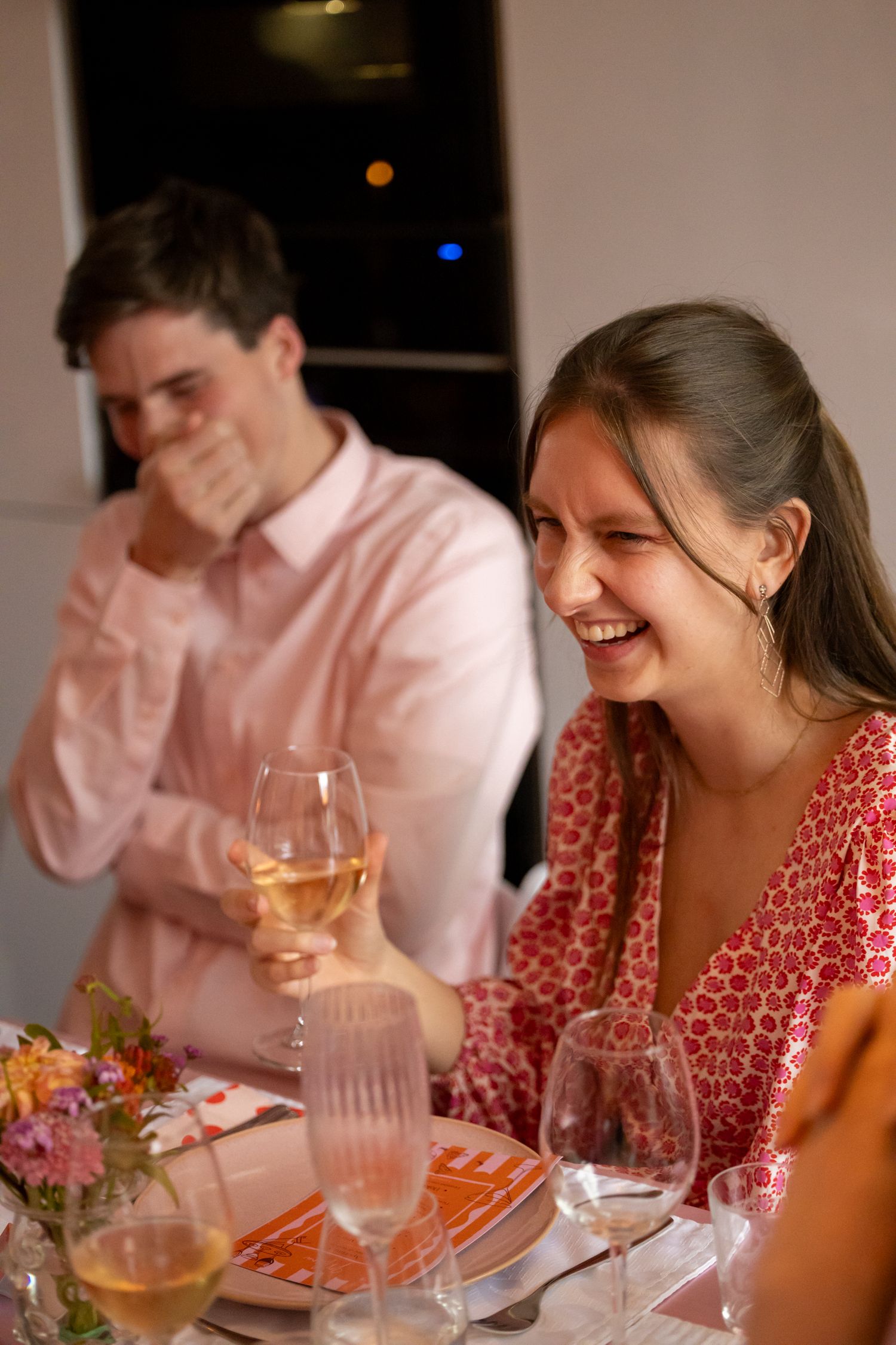 Laughing young woman in red dress holding white wine glass at a warm dinner party gathering.
Evelijn - 21 Diner - Event shoot