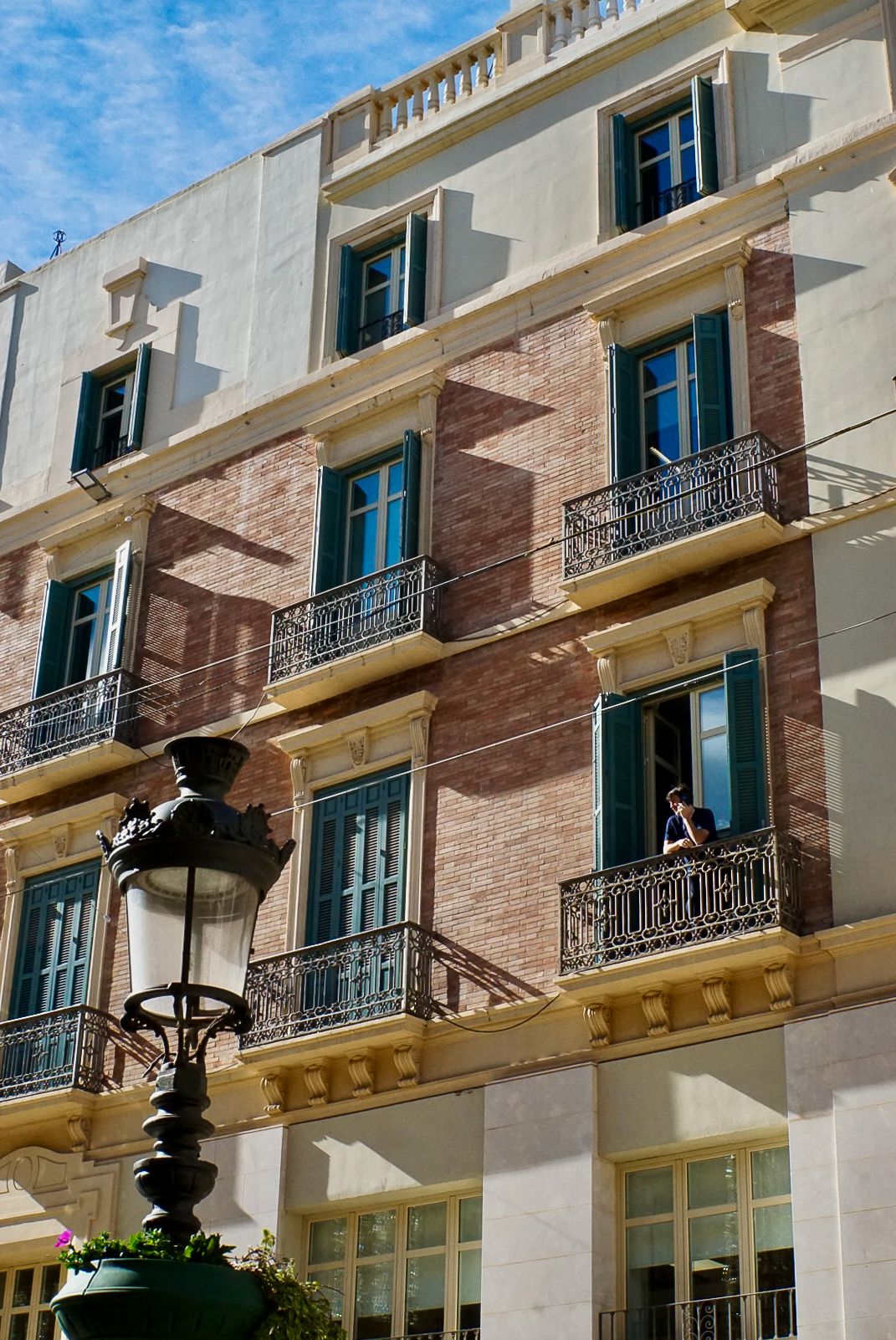 A person stands on an ornate iron balcony of a classic bric building with green shuttered windows and a vintage street lamp.
Malaga
