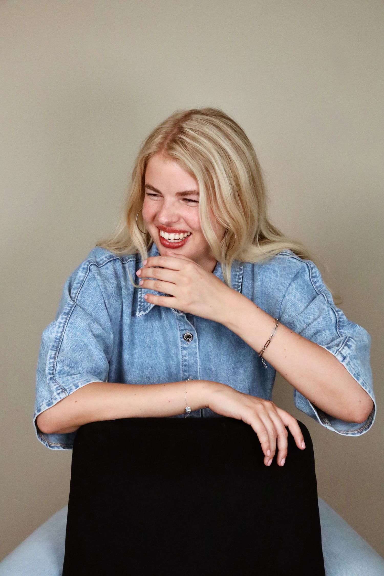 Laughing blonde woman in denim shirt sitting casually, covering mouth with hand, wearing bracelet.
Tess - Soloshoot
