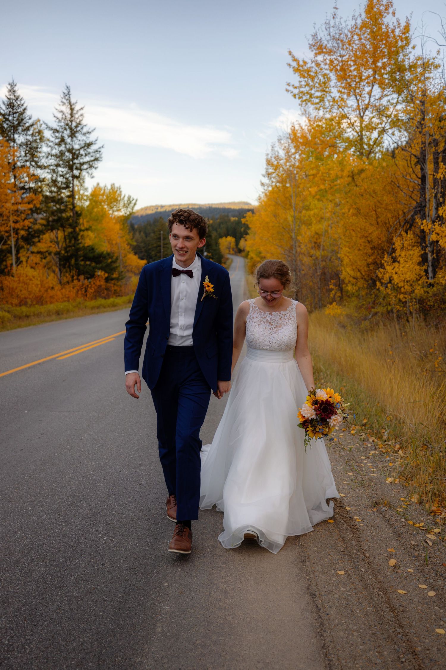 Bride and groom walking along road lined with vibrant golden autumn trees on wedding day.