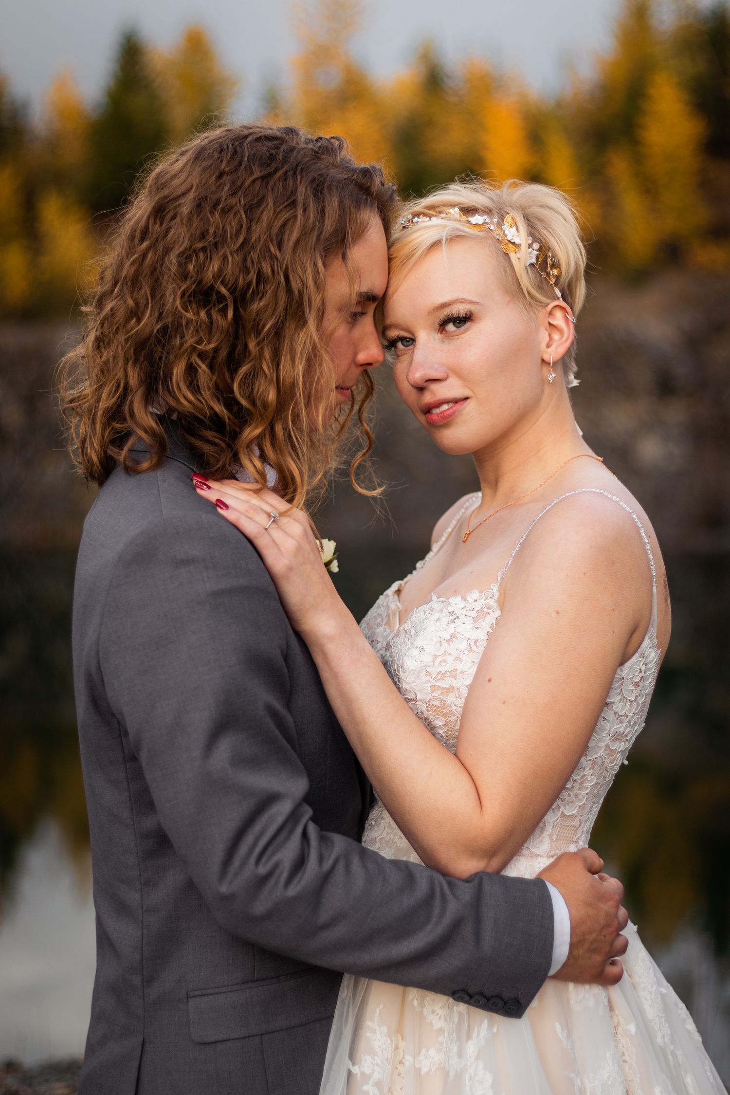 Bride and groom embracing near a lake with autumn foliage in background.