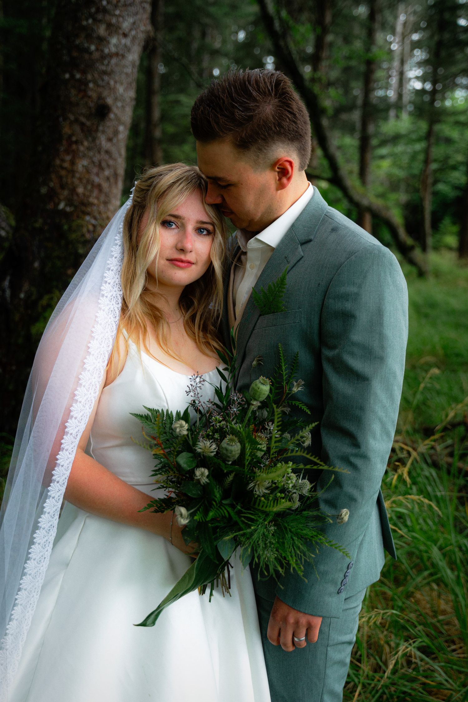 Groom in gray suit tenderly kisses brides head as she holds wild greenery bouquet in dark forest.