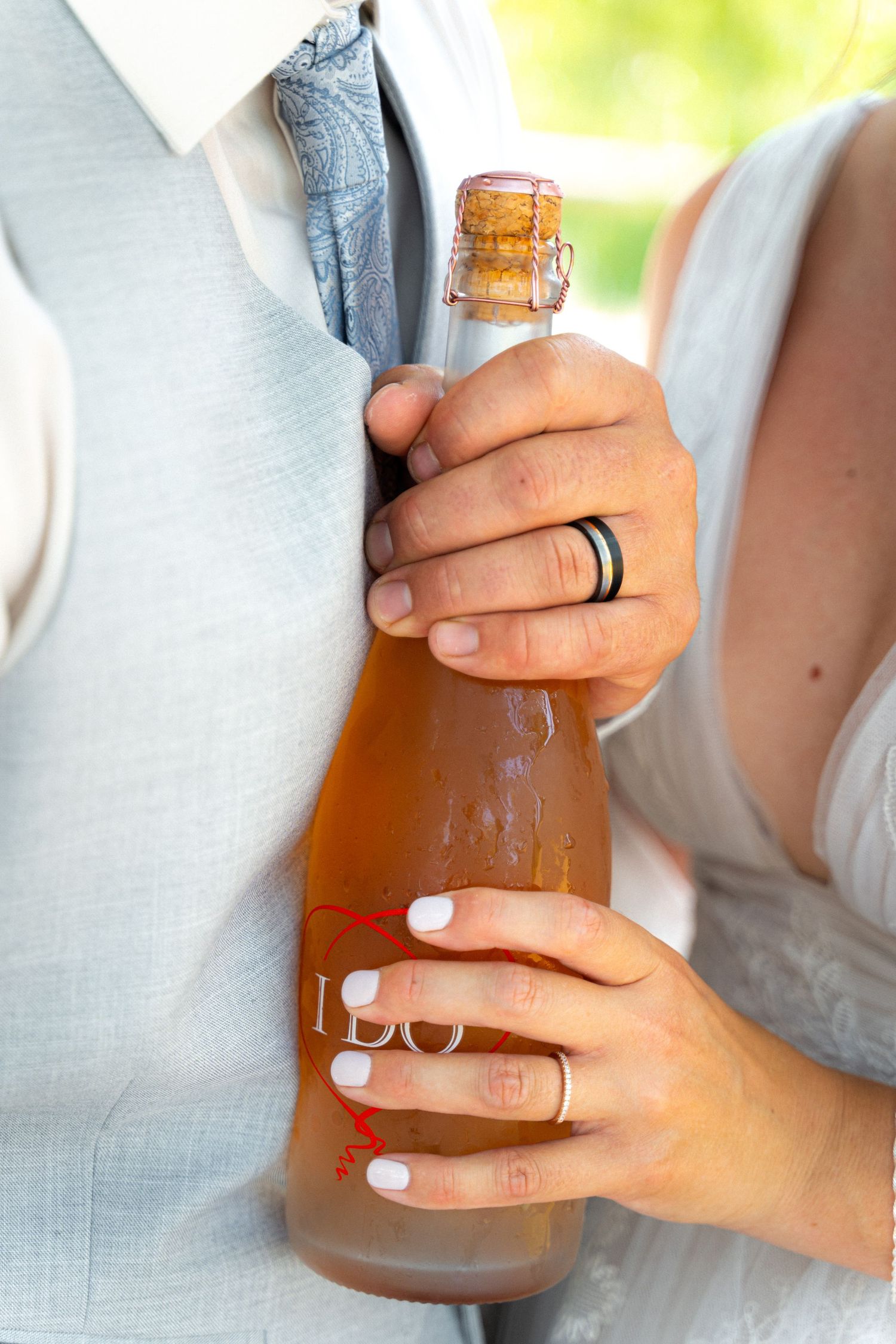 Newlyweds holding a rose champagne bottle with wedding rings visible at outdoor celebration.