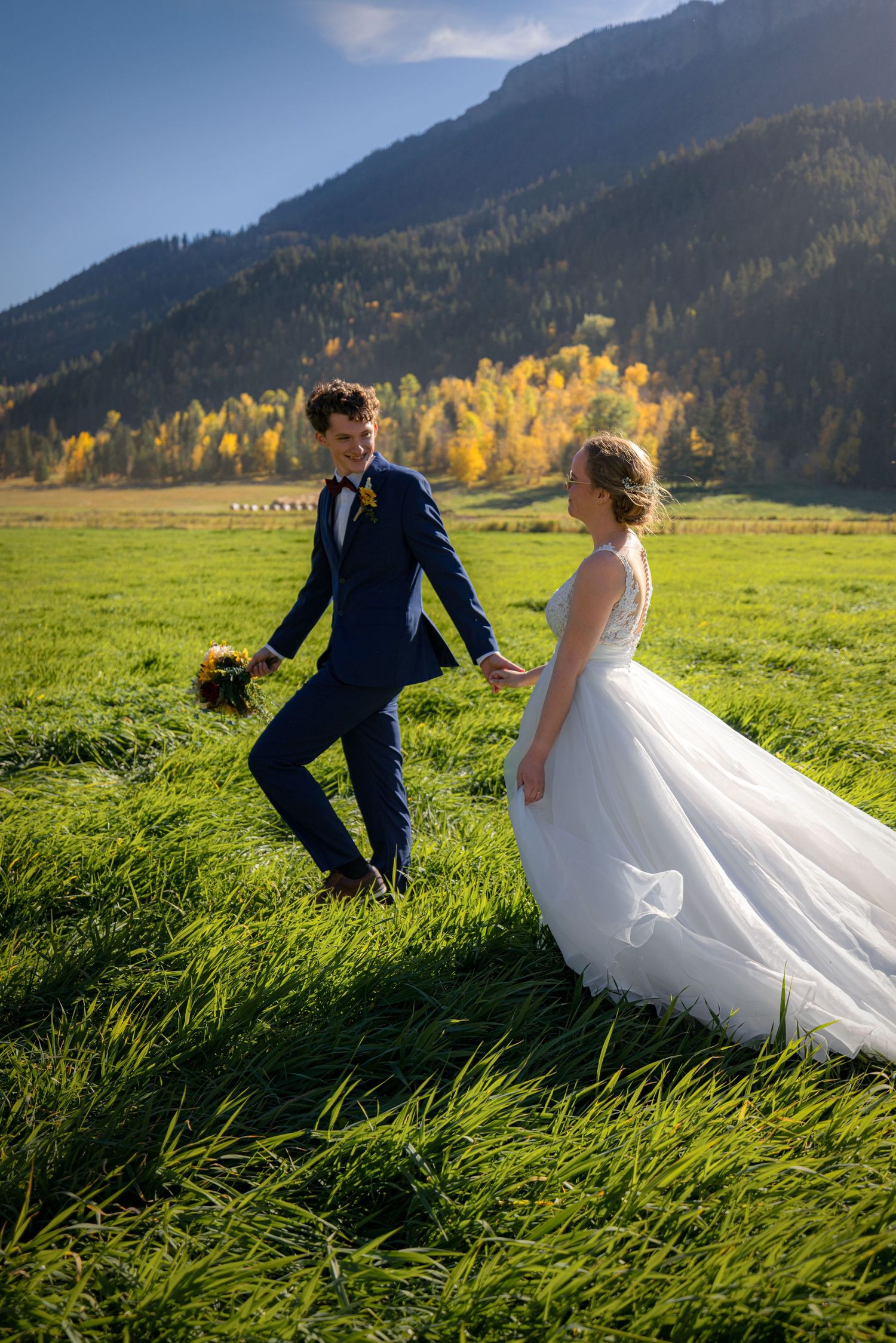Groom in navy suit and bride in white gown dance in green mountain meadow.