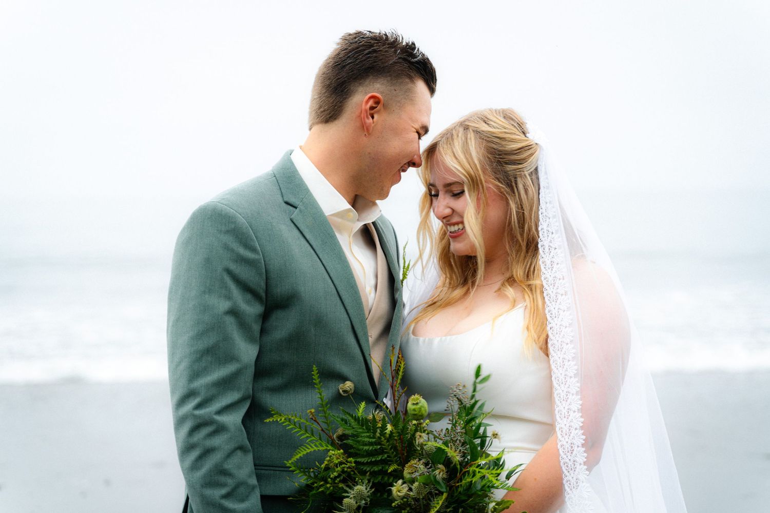 Bride and groom share intimate moment on beach, she holds green bouquet, he wears sage suit, ocean backdrop.