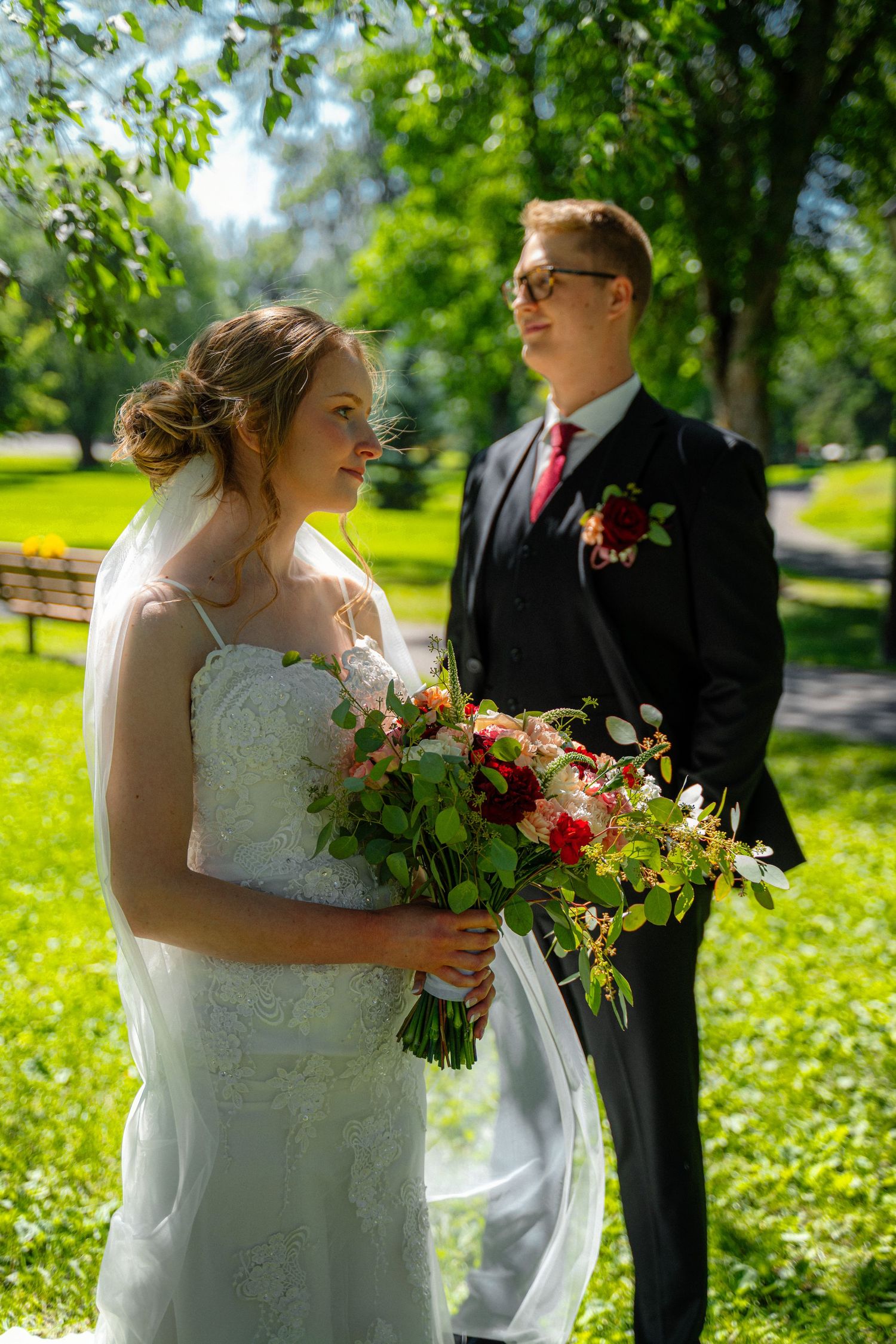 Bride and groom standing in sunny park, bride in white gown holding colorful bouquet.