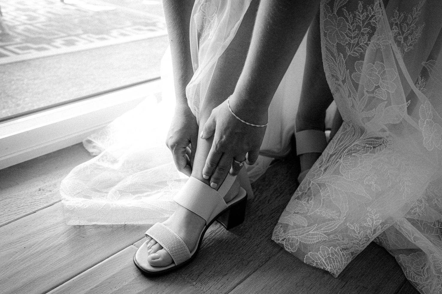 Black and white photo of bride fastening strappy heeled wedding shoes on wooden floor.