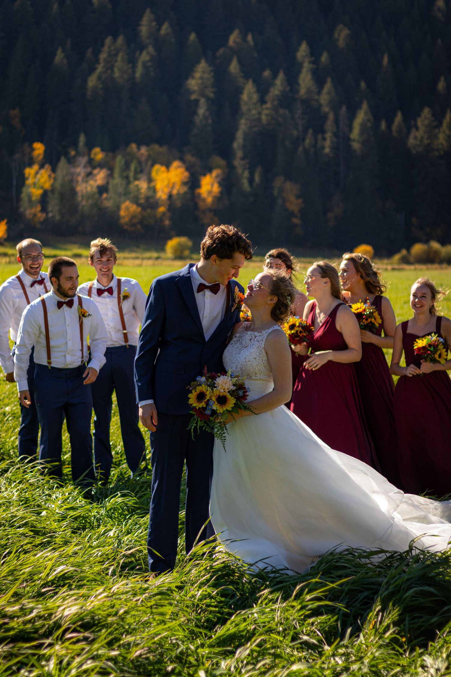 Wedding party posing in sunlit autumn field, bride and groom kissing surrounded by sunflowers and fall foliage.