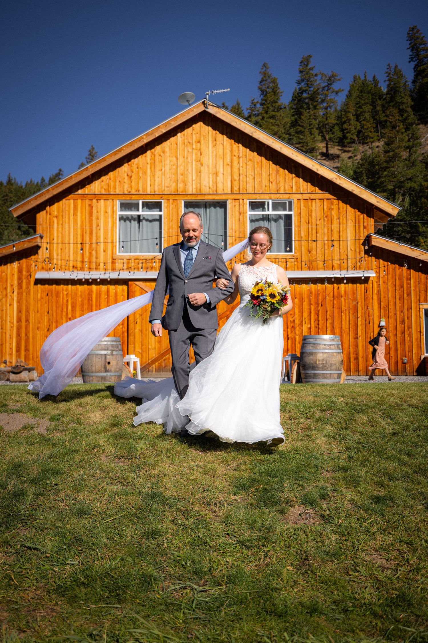 Father walking bride in white gown past a wooden barn, veil flowing in the breeze on a sunny wedding day.