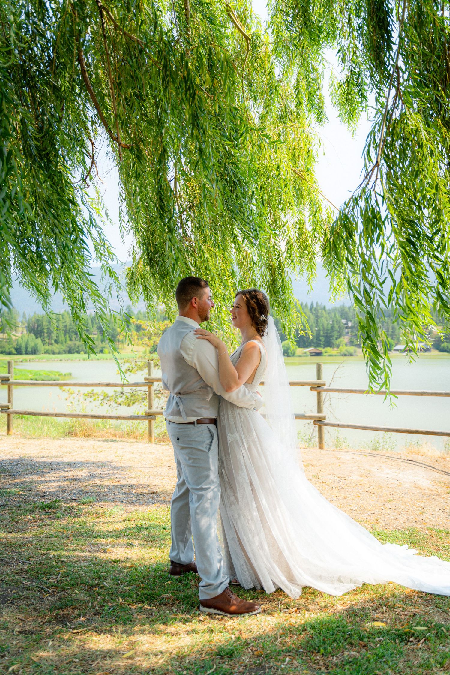 Bride and groom embracing under weeping willow tree by lake on sunny wedding day.