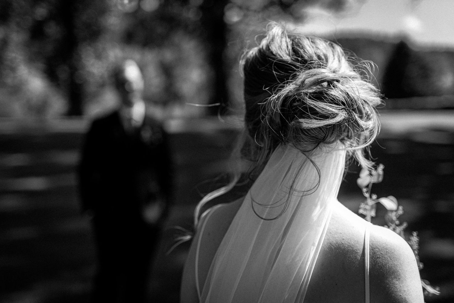 Black and white photo of a bride with an updo veil walking toward a blurred groom in a sunny park.