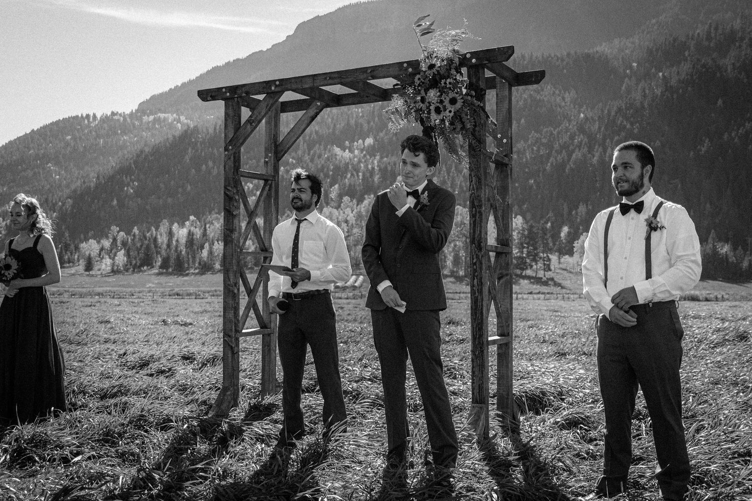 Black and white photo of groomsmen standing at rustic wooden arch with mountain backdrop at outdoor wedding.