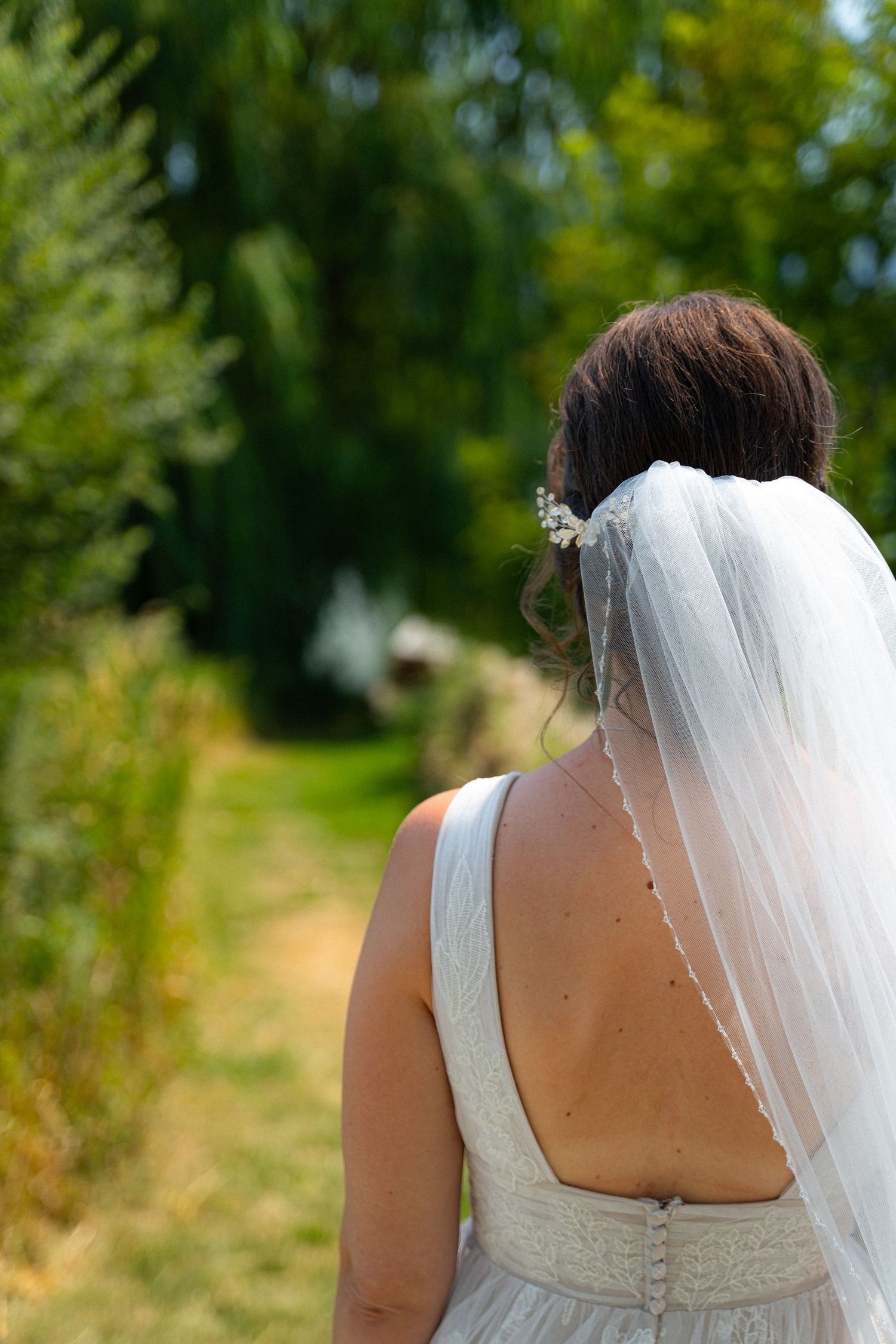 Bride in white lace gown with veil and floral hair accessory walking through lush garden path.