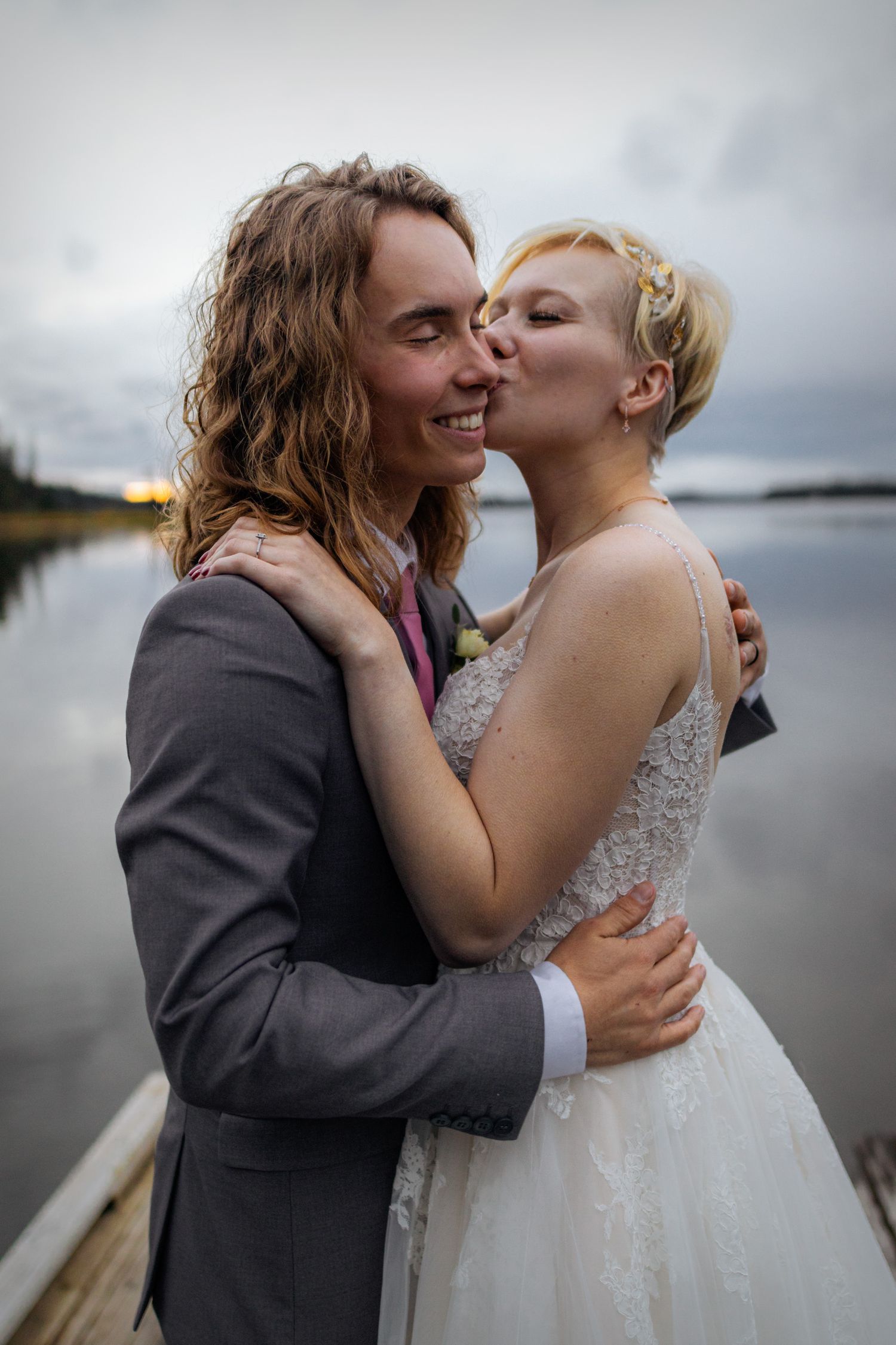 Bride kissing groom on the cheek while embracing near a serene lake at dusk.