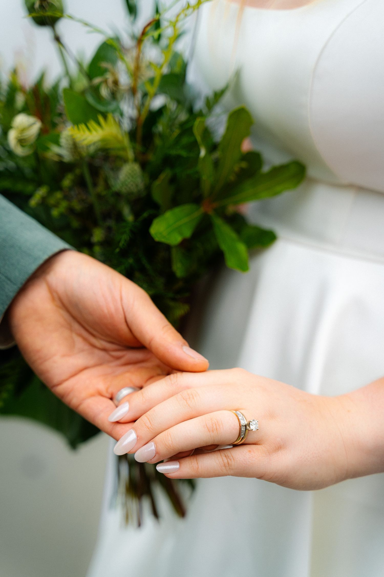 Close-up of bride and groom holding hands with wedding rings and bouquet.