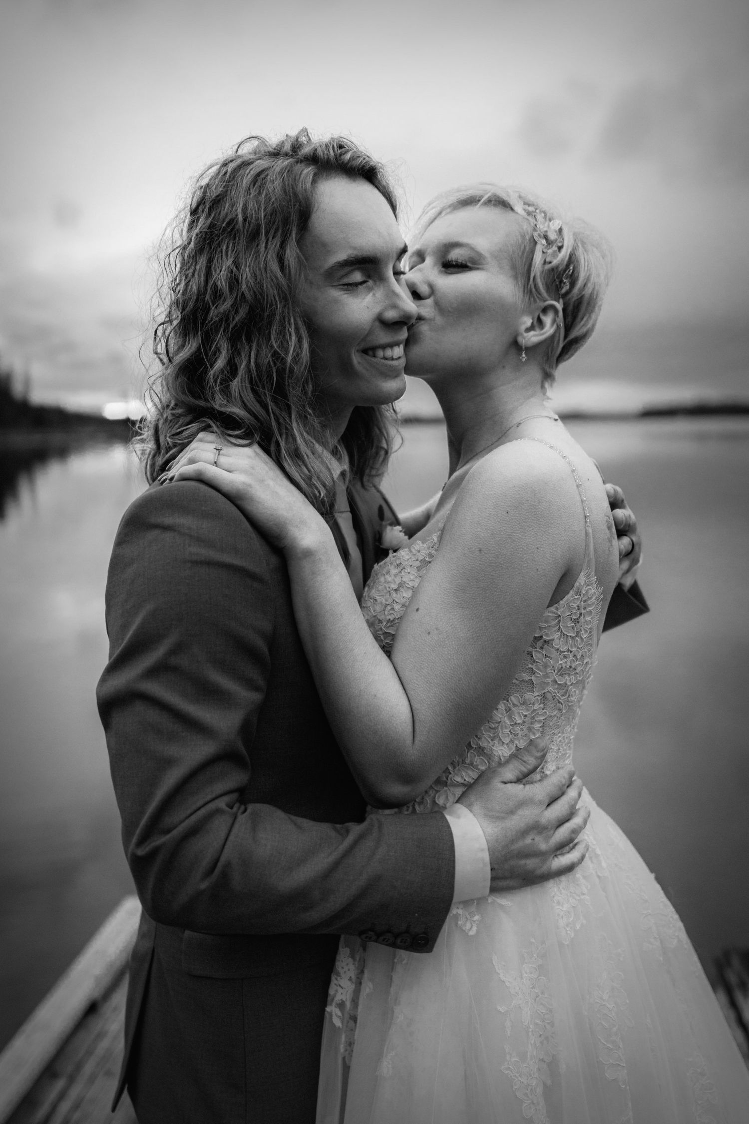 Black and white photo of a bride kissing her smiling groom on the cheek near a calm lake at sunset.