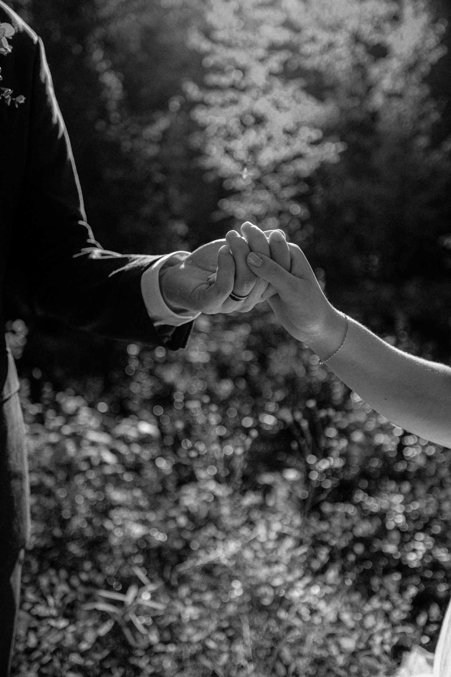 Black and white photo of a couple gently holding hands with bokeh foliage background.