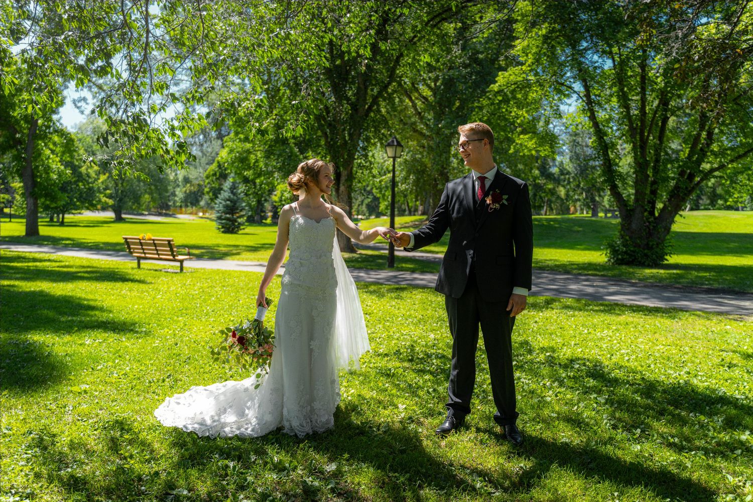 Bride and groom dancing in lush green park, bride in white gown with floral bouquet.