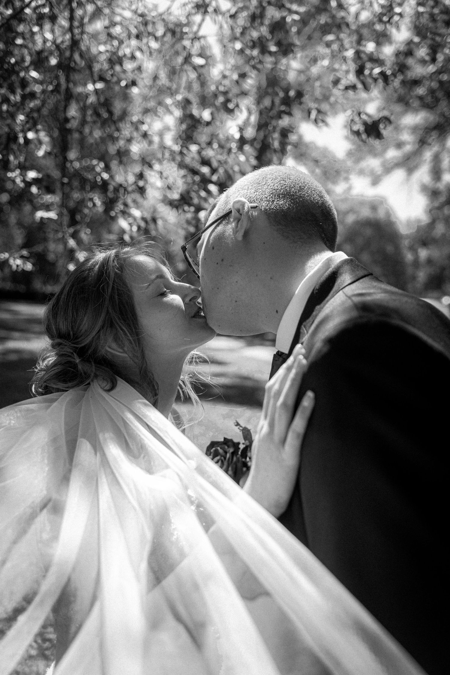 Black and white romantic photo of a bride and groom sharing a kiss under lush trees on their wedding day.