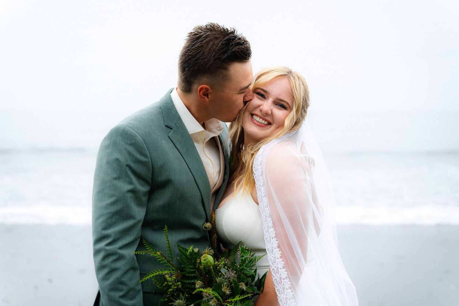 Groom in green suit kissing bride's cheek as she smiles, holding bouquet on beach wedding day.