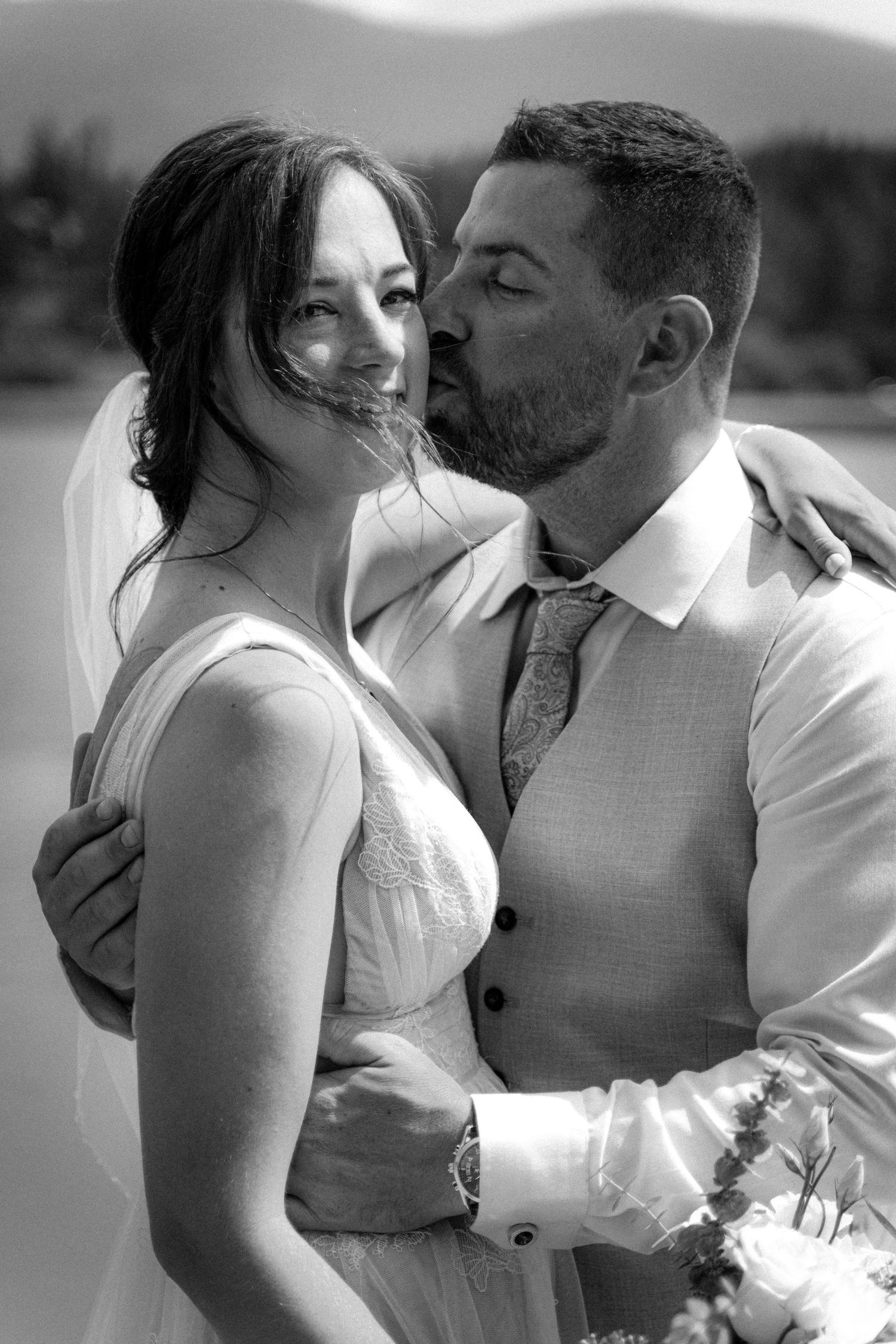 Black and white portrait of groom kissing bride on cheek with mountain lake backdrop.