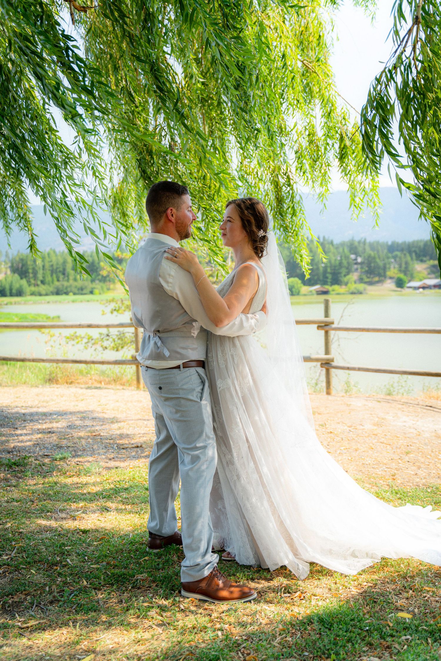 Bride and groom embrace under weeping willow tree by lakeside on sunny wedding day.