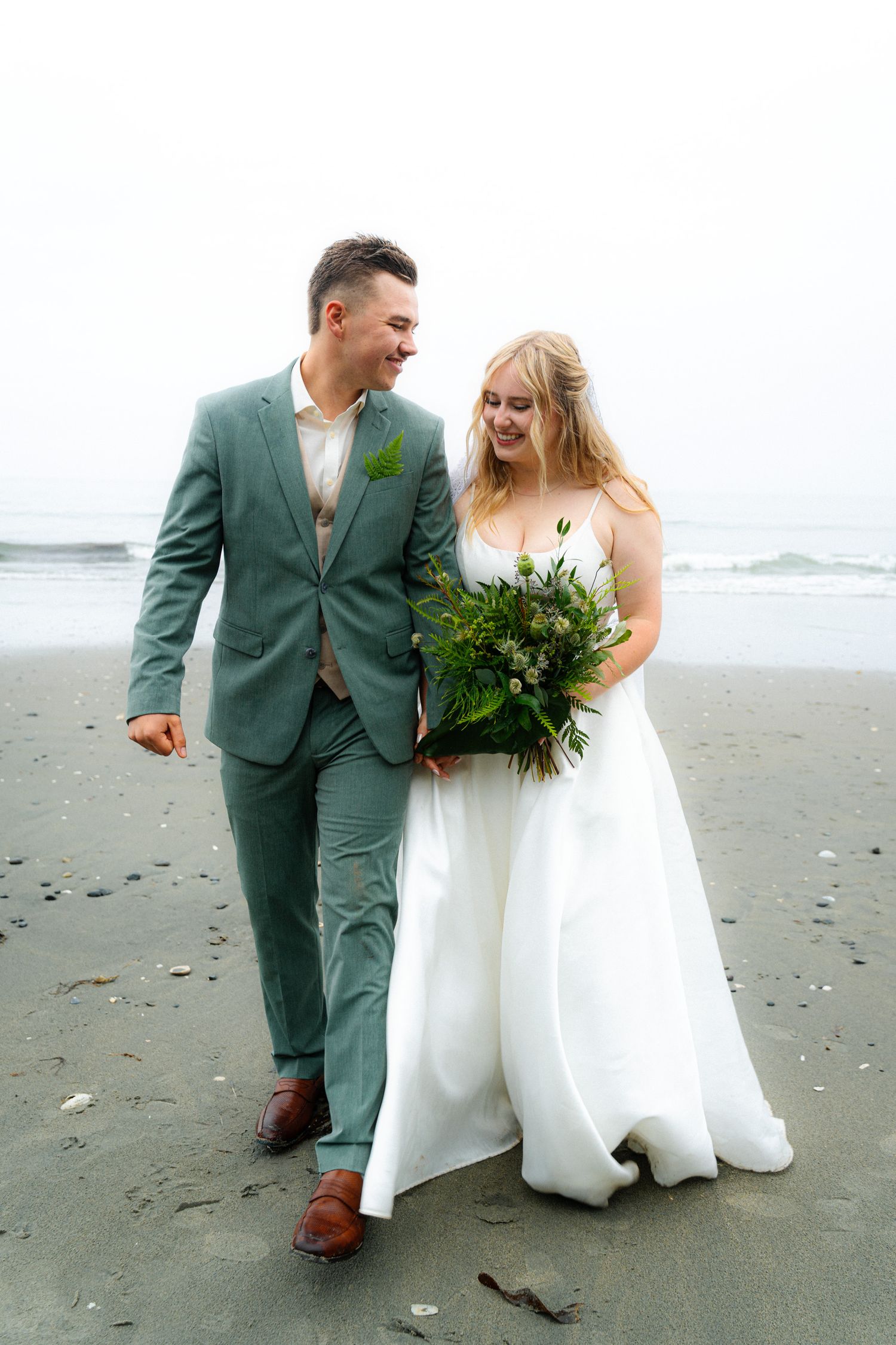Smiling bride and groom walk on misty beach, she in white gown holding green bouquet, he in sage suit.